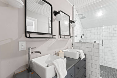 Bright modern farmhouse bathroom with double vessel sinks on a dark gray vanity, black-framed mirrors and sconces, chrome faucets, white towels, and a walk-in subway-tile shower with rainfall showerhead under a beadboard ceiling