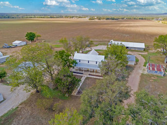 Aerial view of a white two-story country farmhouse with metal roof and wraparound porch, surrounded by trees, outbuildings, a dirt driveway and expansive plowed farmland under a blue sky.