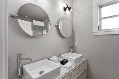Bright modern farmhouse bathroom with double white vessel sinks, round wall mirrors, chrome faucets, distressed wood vanity, subway-tile shower reflected in mirror, and frosted window