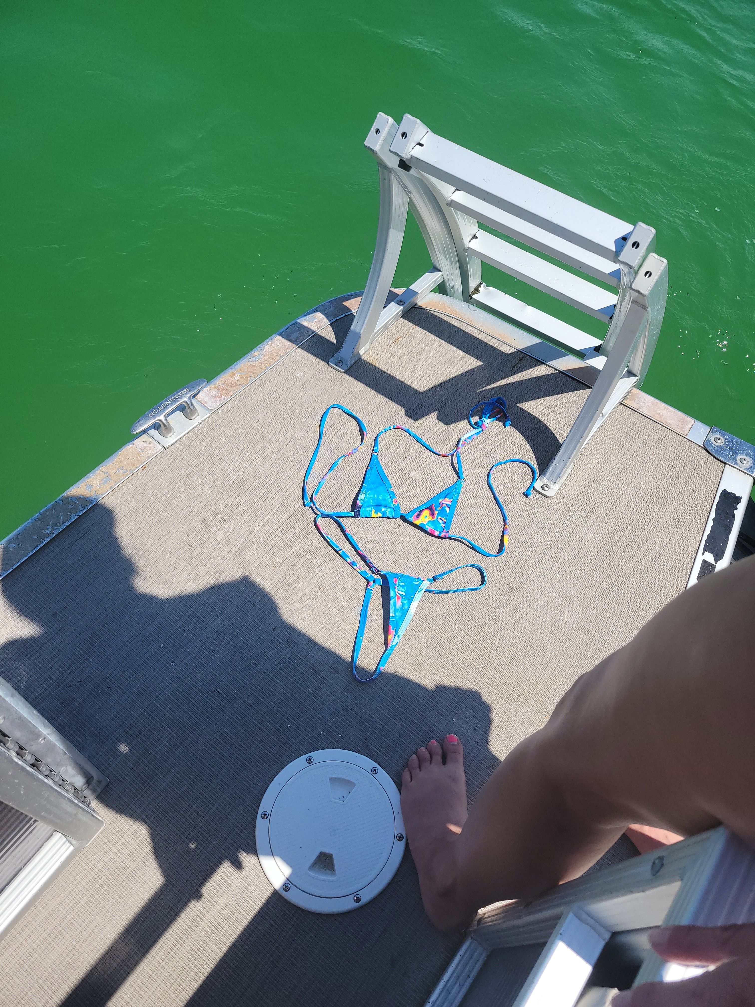 Colorful blue bikini laid on a pontoon boat deck next to a folded swim ladder, bright green lake water and a bare foot with pink-painted toenail — summer boating scene.
