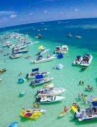 Aerial view of a lively sandbar party with dozens of boats, kayaks, paddleboards, swimmers and inflatables clustered in clear turquoise shallow water under a sunny sky