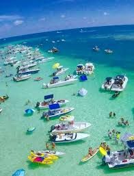 Aerial view of a lively sandbar party with dozens of boats, kayaks, paddleboards, swimmers and inflatables clustered in clear turquoise shallow water under a sunny sky