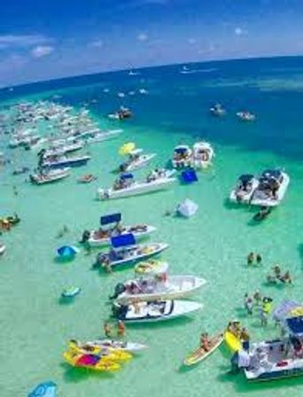 Aerial view of a lively sandbar party with dozens of boats, kayaks, paddleboards, swimmers and inflatables clustered in clear turquoise shallow water under a sunny sky