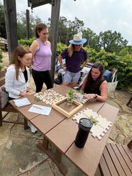 Four women gathered at a wooden picnic table on a stone garden patio, collaborating on a DIY craft session with puzzle-piece mats, beads and natural materials, a jar of herbs and drinks nearby and green shrubs in the background.