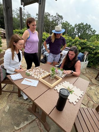 Four women gathered at a wooden picnic table on a stone garden patio, collaborating on a DIY craft session with puzzle-piece mats, beads and natural materials, a jar of herbs and drinks nearby and green shrubs in the background.