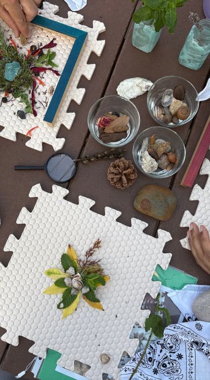 Hands arranging DIY nature collages on white foam puzzle mats at an outdoor wooden table with leaves, flowers, twigs, shells, acorns, pinecone, glass bowls, magnifying glass and blue scissors.