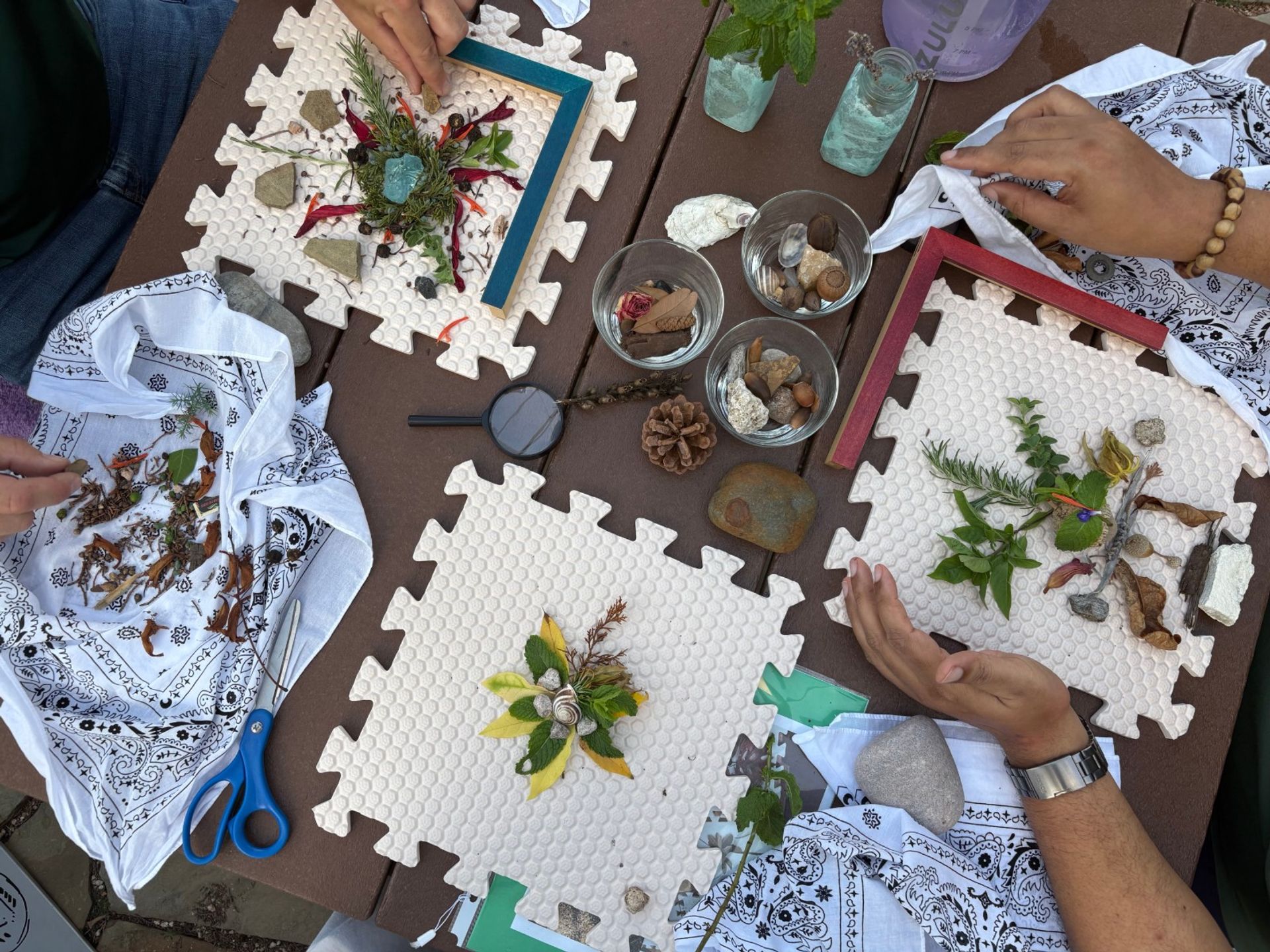 Hands arranging DIY nature collages on white foam puzzle mats at an outdoor wooden table with leaves, flowers, twigs, shells, acorns, pinecone, glass bowls, magnifying glass and blue scissors.