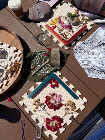 Sunlit patio craft flatlay on a brown outdoor table featuring nature mandala art on foam puzzle tiles with red flower petals, dried seed pods, shells and driftwood, plus bandanas, magnifying glass, glass bowl and takeaway coffee cup.