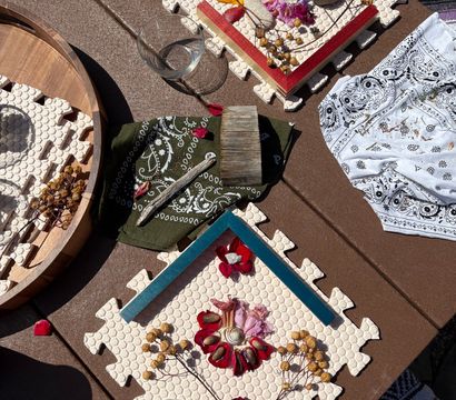 Sunlit patio craft flatlay on a brown outdoor table featuring nature mandala art on foam puzzle tiles with red flower petals, dried seed pods, shells and driftwood, plus bandanas, magnifying glass, glass bowl and takeaway coffee cup.