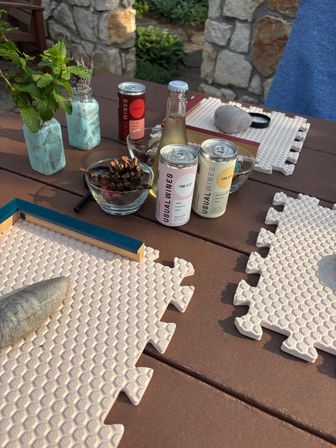 Sunlit backyard patio table with canned wine and a small bottled wine, glass bowls of pinecones, two turquoise jars of mint and lavender, a smooth stone, book and interlocking foam placemats — casual outdoor table decor.