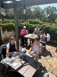 Sun-dappled stone patio with five women seated under a wooden pergola, crafting flower and plant arrangements at wooden tables surrounded by green hedges.