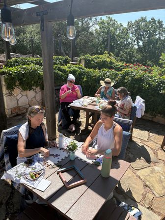 Sun-dappled stone patio with five women seated under a wooden pergola, crafting flower and plant arrangements at wooden tables surrounded by green hedges.