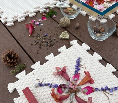 Outdoor garden craft scene: nature mandala of pink and purple flower petals, seeds, acorns and a pinecone arranged on white foam puzzle tiles on a wooden table.