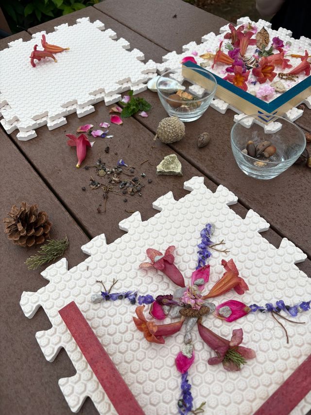 Outdoor garden craft scene: nature mandala of pink and purple flower petals, seeds, acorns and a pinecone arranged on white foam puzzle tiles on a wooden table.