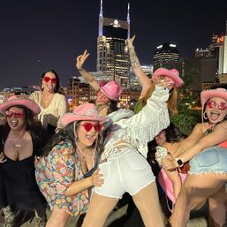 Group of friends in pink cowgirl hats and heart-shaped sunglasses posing playfully at night with downtown Nashville skyline and an illuminated twin-spired tower in the background.