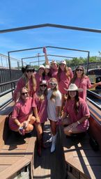 Ten friends in matching dusty-rose shirts and cowboy hats riding an open-air wooden wagon, holding drinks and wearing boots under a bright blue sky — fun country-style daytime outing
