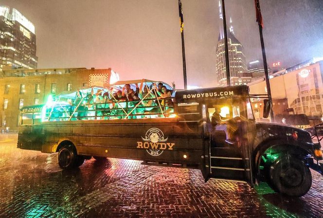 Neon-lit open-air party bus full of cheering passengers on a rainy night in downtown Nashville, wet brick street and illuminated skyline