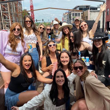 Group of friends enjoying a sunny rooftop street-party in a downtown entertainment district, smiling and holding canned drinks while wearing sunglasses, hats and casual summer outfits with city buildings and a construction crane in the background