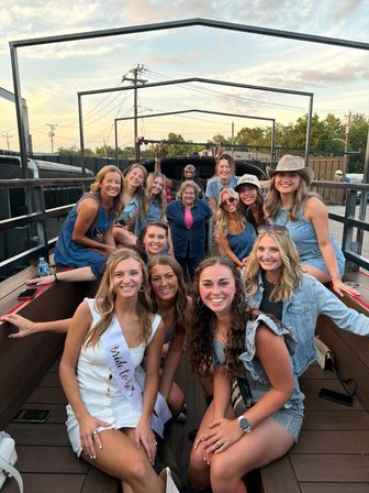 Group of smiling women in casual summer outfits on an open-air party bus trailer celebrating a bachelorette at sunset