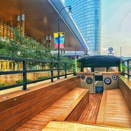 Open-air wooden-deck riverboat with bench seating docked along a modern downtown waterfront, glass skyscraper and covered promenade with trees and colorful banners in the background.
