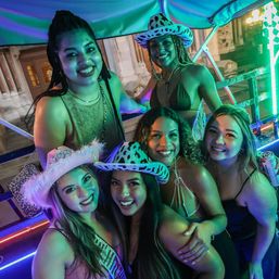 Six smiling friends on a neon-lit outdoor party ride in a city at night, wearing swimsuits, cowgirl and fuzzy hats, close-up group selfie with colorful green and purple lights