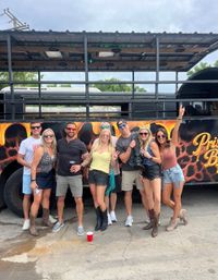 Eight friends posing outdoors in front of a decorated party bus, smiling in casual summer outfits with sunglasses, cowboy boots and drinks.