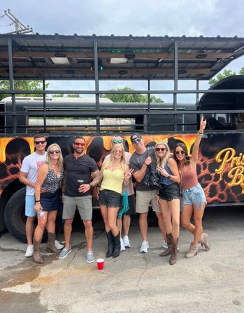 Eight friends posing outdoors in front of a decorated party bus, smiling in casual summer outfits with sunglasses, cowboy boots and drinks.