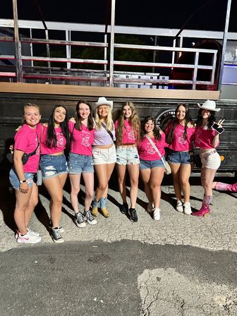 Eight women in matching pink shirts and denim shorts posing at night in front of a parked trailer, some wearing cowboy hats and boots — outdoor country-night group celebration.