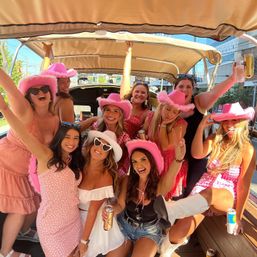 Group of women wearing pink cowboy hats and summery outfits smiling and holding drinks on a covered party cart on a sunny city street — bachelorette/girls' weekend celebration.