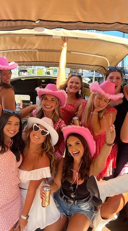 Group of women wearing pink cowboy hats and summery outfits smiling and holding drinks on a covered party cart on a sunny city street — bachelorette/girls' weekend celebration.