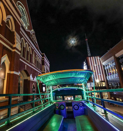 Neon-lit open-top party bus deck on a downtown street at night, green LED benches under a canopy with the moon above and a historic red-brick theater and illuminated modern buildings in the background.