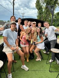 Smiling group of friends at an outdoor summer party on artificial turf, holding drinks on a deck with stools, trees and a dramatic cloudy sky.