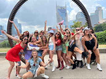 Large group of friends in summer outfits and cowboy boots posing and cheering under a circular metal sculpture with the Nashville skyline and twin‑spired tower in the background
