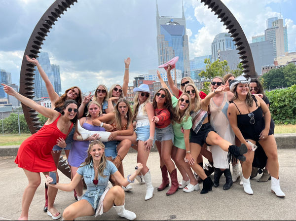 Large group of friends in summer outfits and cowboy boots posing and cheering under a circular metal sculpture with the Nashville skyline and twin‑spired tower in the background