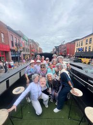 Group of women in gray wigs and playful outfits posing and celebrating on an open-top party vehicle on a busy downtown street lined with brick buildings and neon bar signs under an overcast sky