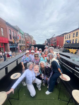 Group of women in gray wigs and playful outfits posing and celebrating on an open-top party vehicle on a busy downtown street lined with brick buildings and neon bar signs under an overcast sky