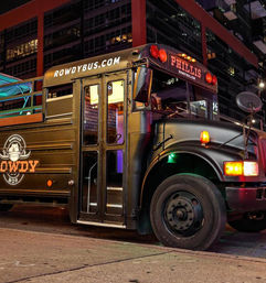 Sleek black party bus parked on a downtown city street at night, folding entrance open to a lit interior, rooftop deck with railings and colorful lights.