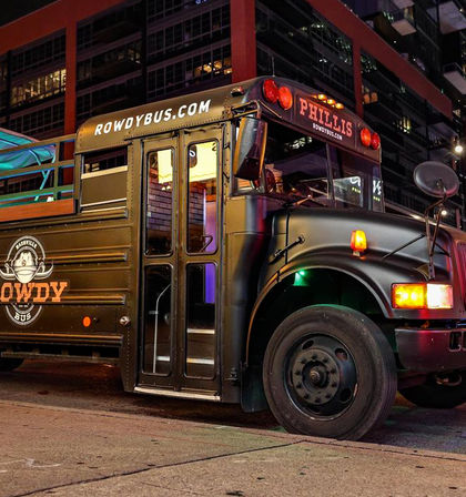 Sleek black party bus parked on a downtown city street at night, folding entrance open to a lit interior, rooftop deck with railings and colorful lights.