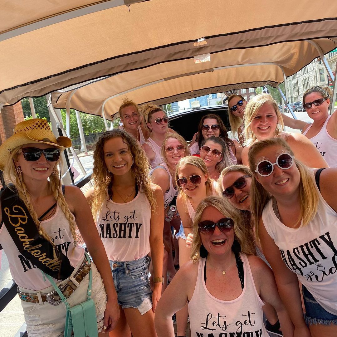 Smiling bachelorette group on a covered city party cart, wearing matching 'Let’s get NASH TY' tank tops, sunglasses and casual summer outfits for a downtown celebration