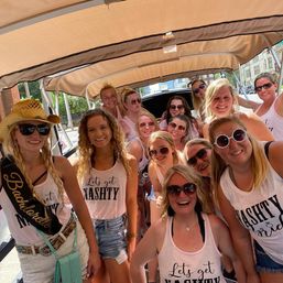 Smiling bachelorette group on a covered city party cart, wearing matching 'Let’s get NASH TY' tank tops, sunglasses and casual summer outfits for a downtown celebration