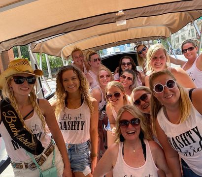 Smiling bachelorette group on a covered city party cart, wearing matching 'Let’s get NASH TY' tank tops, sunglasses and casual summer outfits for a downtown celebration