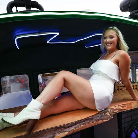 Smiling woman in a white dress and ankle boots lounging on a live-edge wooden bar under blue neon lights inside a mobile bar.
