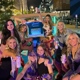 Group of eight smiling women on a neon-lit party trolley parked on a downtown street at night, raising canned drinks and posing in dresses for a lively girls' night out.