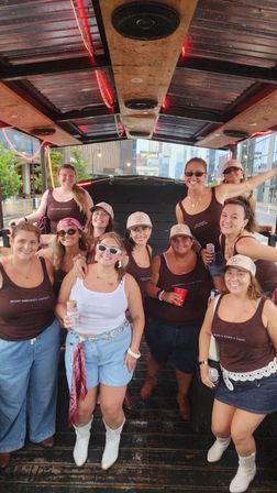 Smiling group of women on a covered party bike in a downtown street, wearing matching tank tops, cowboy boots and caps and holding drinks — festive bachelorette-style city celebration.