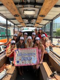 Smiling group of women on a wooden party trolley on a sunny downtown street, wearing matching trucker hats and holding a red-check bachelorette banner that reads All American Bride during an urban celebration