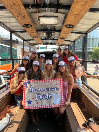 Smiling group of women on a wooden party trolley on a sunny downtown street, wearing matching trucker hats and holding a red-check bachelorette banner that reads All American Bride during an urban celebration