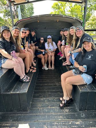 Bachelorette group of women seated on a black wooden party wagon, many wearing matching black shirts and caps and one woman in white at center, holding canned drinks on a sunny outdoor, tree-lined day