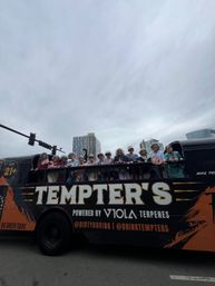 Group of friends on an open-top party bus waving from the upper deck on a cloudy downtown street with high-rise buildings in the background