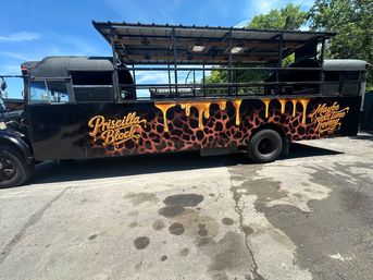 Black converted party bus parked in a sunny lot with open-sided roof, leopard-print lower panels and golden honey-drip mural accents over stylized script lettering — custom exterior art on a retro bus.
