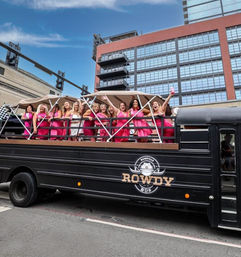 Group of women—mostly in bright pink dresses with one in white—cheering on an open-top party bus rolling through a downtown city street under a blue sky.